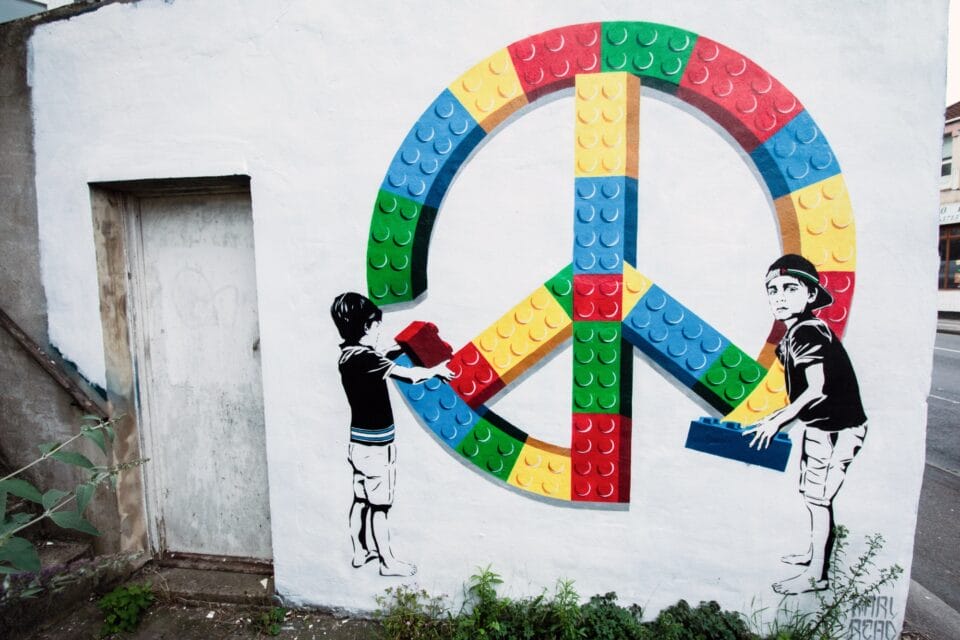 a street art mural of a peace sign made out of colorful LEGO bricks, with two kids assembling it